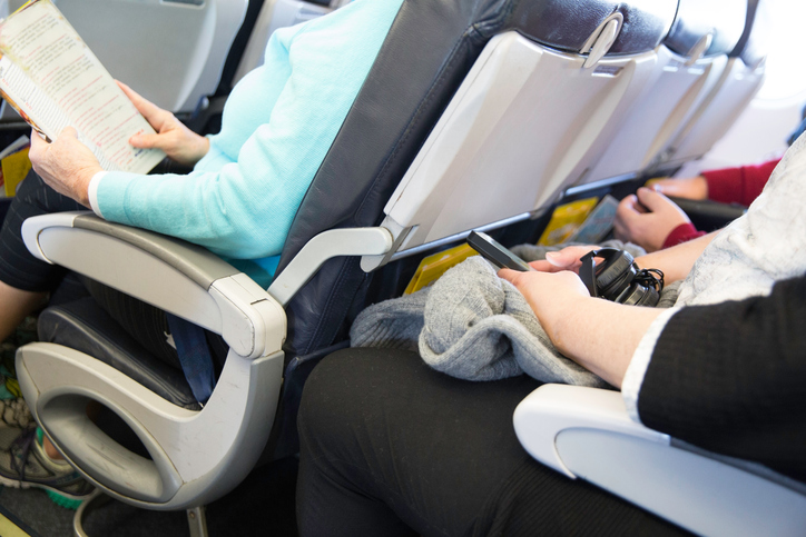 In this real situation a row of unidentifiable passengers are sitting in their seats on an airplane. The seating is tight. Their legs are touching the seats in front of them.